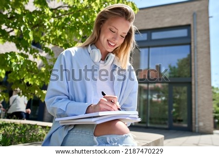 Pretty happy smiling girl university student holding notebook studying writing checklist sitting outdoors, taking notes learning college academic educational course concept. Royalty-Free Stock Photo #2187470229