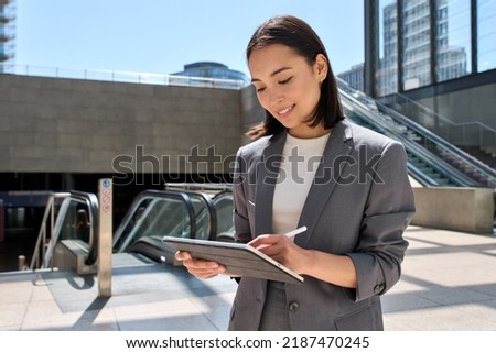 Young Asian woman entrepreneur standing in subway using digital tablet modern device. Smiling busy korean businesswoman manager wearing suit holding pad computer remote working online outdoor.