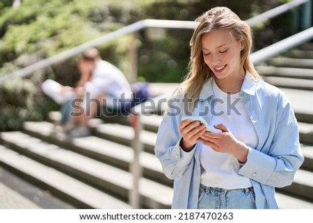 Young smiling woman university student holding cell phone using mobile apps technology on smartphone looking at cellphone texting in messengers writing messages standing outside campus. Royalty-Free Stock Photo #2187470263
