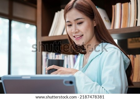 Female student is drinking coffee and reading literature on tablet while sitting to take a break after studying online learning in library.