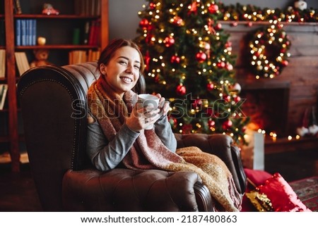 Young Woman Sits in an Armchair at Home Against Background of Christmas Tree and Fireplace. Hold Cup of Hot Drink in Hands. Merry Christmas and Happy New Year greeting card Royalty-Free Stock Photo #2187480863