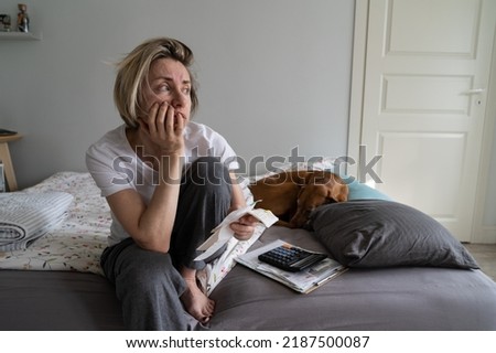 Depressed middle-aged woman looks away to get distracted from financial crisis and life problems. Unkempt mature female props chin with hand holding unpaid bills on bed regretting about past closeup Royalty-Free Stock Photo #2187500087