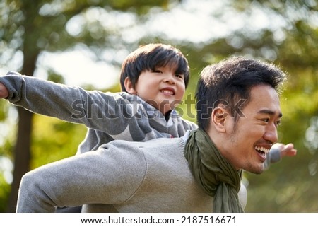 young asian father carrying son on back having fun enjoying nature outdoors in park Royalty-Free Stock Photo #2187516671