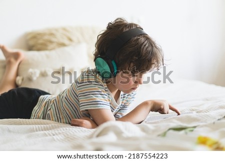 Cute boy lying on the bed at home and playing with pc tablet or reading online and listening to music with wireless headphones