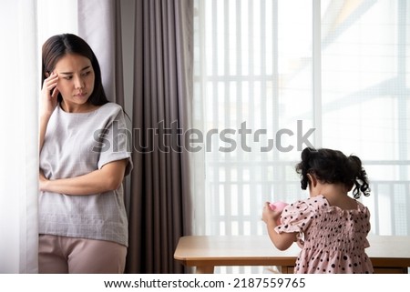 Unhappy Asian mother touching forehead, feeling tired of bad daughter's behavior at home. Offended little child girl sitting on different side on table. stubborn and mischievous behavior