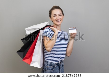 Portrait of smiling positive woman in striped T-shirt holds and showing paper bags and credit card, contactless payments, easy shopping with debit card. Indoor studio shot isolated on gray background.