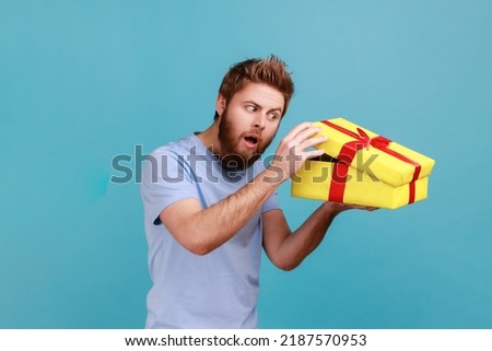 Portrait of cunning handsome bearded man unboxing yellow gift box, looking inside with curious facial expression, birthday present. Indoor studio shot isolated on blue background. Royalty-Free Stock Photo #2187570953