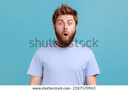 Portrait of shocked bearded man gasps from wonder, holds breath, stares at something horrible keeps mouth opened, being astonished. Indoor studio shot isolated on blue background.
