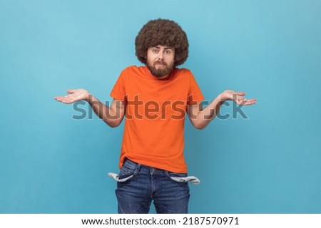 Portrait of man with Afro hairstyle wearing orange T-shirt showing empty pockets and looking frustrated about loans and debts, has no money, jobless. Indoor studio shot isolated on blue background. Royalty-Free Stock Photo #2187570971