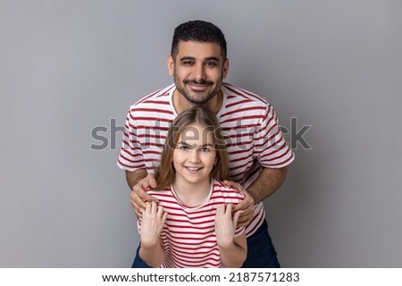 Portrait of delighted smiling father and daughter in striped T-shirts standing and looking at camera, dad embracing little kid, expressing happiness. Indoor studio shot isolated on gray background.