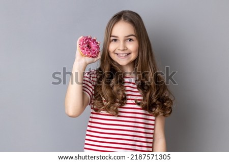 Portrait of little girl wearing striped T-shirt standing holding doughnut and smiling at camera, showing sweet sugary confectionary. Indoor studio shot isolated on gray background. Royalty-Free Stock Photo #2187571305