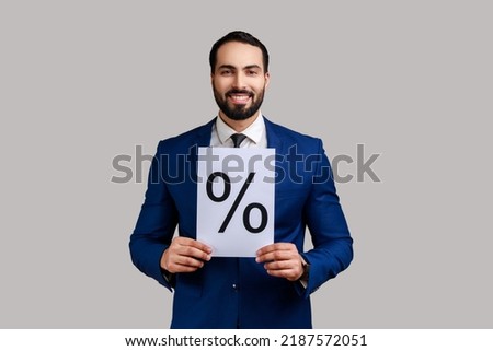 Positive smiling bearded man holding showing paper with percent sign inscription, looking at camera, wearing official style suit. Indoor studio shot isolated on gray background.