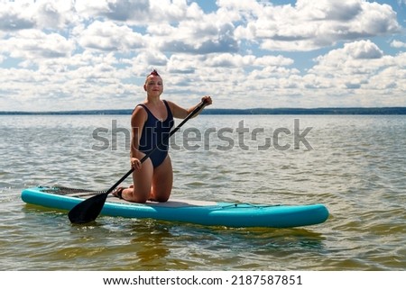 a woman in a closed swimsuit with kneeling on a SUP board with a paddle floats on the water against the blue sky. Horizontal photo