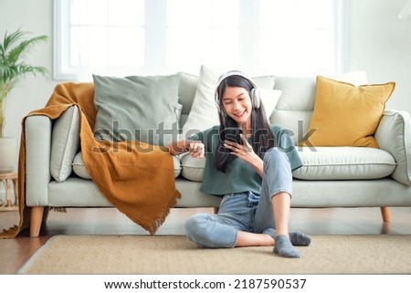 Happy asian woman listening to music from mobile phone while sitting on the rug beside to the sofa at homes, Smiling girl relaxing with headphones in morning, Time to relax. Royalty-Free Stock Photo #2187590537