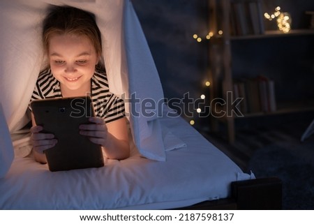 Close up of little girl watching cartoons on the digital tablet at night under blanket, copy space. blue light has a negative effect on child's eyes.