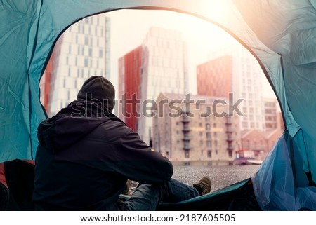 View from inside of a homeless tent. Man in dark dirty cloths sitting by the entrance looking at rich high value district houses. Living during financial crisis concept. Dreaming on better future. Royalty-Free Stock Photo #2187605505