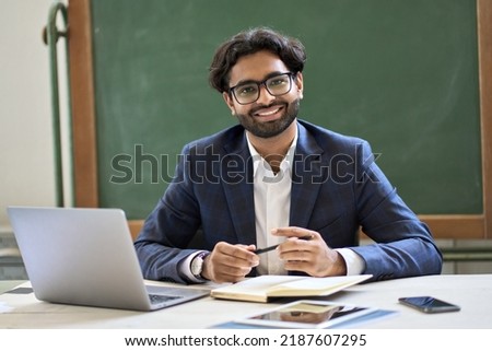 Happy young indian arabic businessman professional coach, teacher or university professor wearing suit looking at camera sitting at work desk in classroom office posing for portrait at workplace. Royalty-Free Stock Photo #2187607295