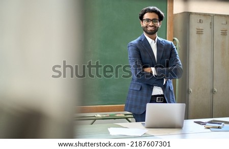 Happy young adult indian business man coach wearing suit looking at camera standing in office. Arab college teacher or university professor posing for portrait at workplace desk in classroom. Royalty-Free Stock Photo #2187607301