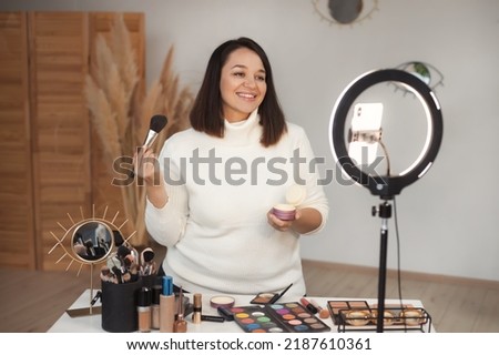Plus size latin woman working as make up artist and beauty blogger recording video on mobile phone at beauty salon with led ring lamp