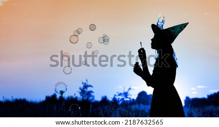 Black silhouette of witch blowing soap bubbles isolated on fairy sunset sky background.Beautiful young woman in black robe and wizard hat conjuring, making magic. Halloween party art design.Copy space