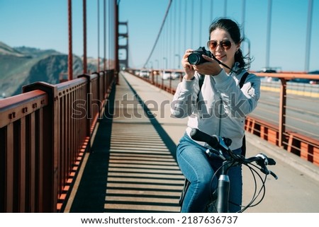 portrait Asian female tourist wearing sunglasses is shooting a photo with a digital camera in the sun on bike at golden gate bridge in San Francisco California