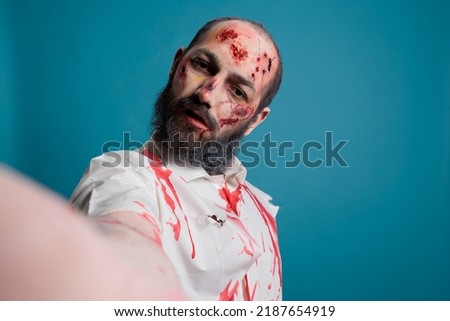 Undead zombie holding camera to take photo, looking creepy and dangerous over blue background. Scary brain eating devil with horror look and aggressive bloody face taking frightening picture.