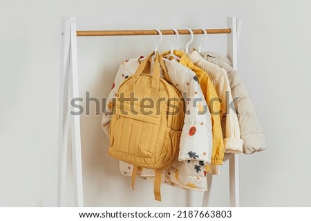 Wooden Clothing Rack with children's autumn outfit. Yellow backpack,  jacket and sweaters on hangers in wardrobe. Nursery Storage Ideas. Home kids wardrobe. 