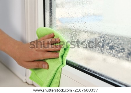 Woman wiping window glass with drops of condensate indoors, closeup Royalty-Free Stock Photo #2187680339