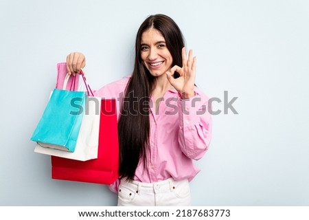 Young caucasian woman going to buy some clothes isolated on blue background cheerful and confident showing ok gesture.