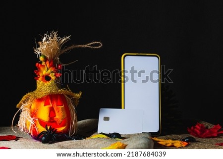 Credit card on a cell phone with a yellow case and the screen on, both standing next to an ornate Halloween pumpkin, on sackcloth and surrounded by toy spiders and fallen leaves next to pine cones.