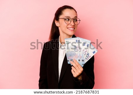Young business caucasian woman holding bank notes isolated on pink background looks aside smiling, cheerful and pleasant.