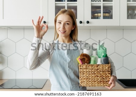 Housemaid from cleaning services holding a box with detergents and household chemical products, showing ok sign, standing on kitchen, looking at camera. Easy and quick housework advertising concept