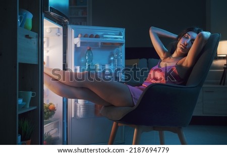 Exhausted woman suffering from the heat during the summer heatwave, she is sitting in front of the open fridge and cooling herself Royalty-Free Stock Photo #2187694779