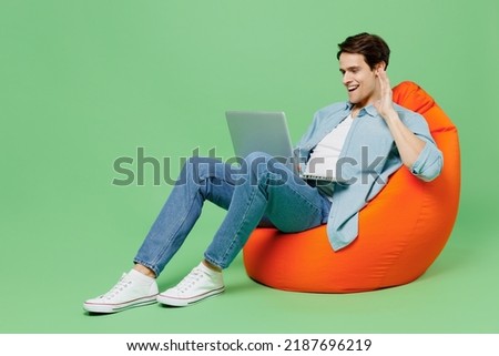 Full size young brunet man 20s wears blue shirt sit in bag chair get video call use laptop pc computer conducte pleasant conversation greet with hand isolated on plain green background studio portrait