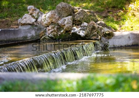Close up urtificial creek water runs off. Mountain rapids with a stream of water in a river. Little mountain waterfall, stones and green grass in public park, forest, nature reserve. Natural landscape Royalty-Free Stock Photo #2187697775