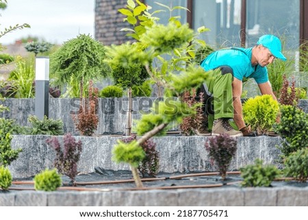 Caucasian Gardener in Blue Cap and Shirt Arranging Newly Created Front Yard Garden by Planting Various Plants in the Soil Covered with Weed Membrane. Professional Landscape Design Theme. Royalty-Free Stock Photo #2187705471