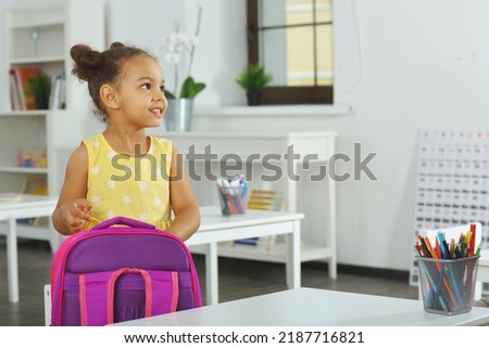 an African-American girl puts a notebook in a school backpack. A child in a yellow dress stands in the classroom and prepares for a lesson
