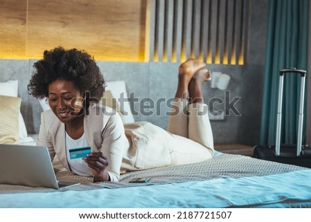 The woman lies on the bed with a laptop and a credit card and enters the data for the purchase. An African business woman makes online payments from a hotel room.