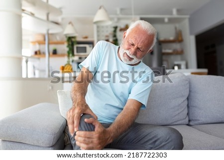 The older man is sitting on the couch at home, has pain in the knee joint, holding his leg, osteoarthritis concept. Royalty-Free Stock Photo #2187722303