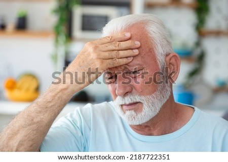 Tired, depressed senior man sitting on couch in living room feeling hurt and lonely. Aged, white-haired man touching forehead suffering from severe headache or recalling bad memories Royalty-Free Stock Photo #2187722351