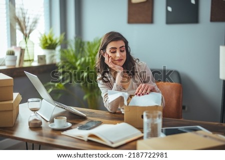 The surprised facial expressions of the girl sitting at the table in the living room open a large cardboard box and unpack the ordered goods via the internet website, she feels happy and amazed 