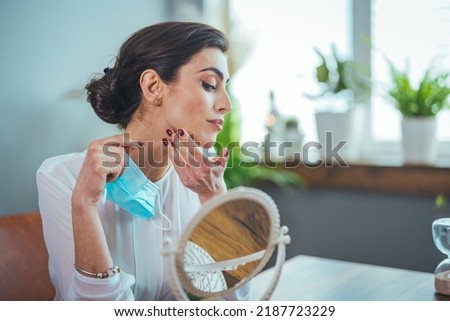 Young beautiful woman looking herself in the mirror at home. Worried about acne caused by wearing a mask. Maskne. Problems with acne. Skin allergy From wearing a mask