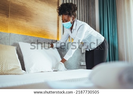 Afro maid working at a hotel doing room disinfection wearing a face mask in time of COVID-19 pandemic. Female maid with medical mask changing bed linen on the bed