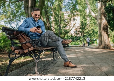 Smiling businessman text messaging on smart phone. Confident young male professional is networking while sitting on bench. He is wearing formals at the park in the city. Royalty-Free Stock Photo #2187742213