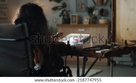 Back view of a woman in a wheelchair with physical disability painting with watercolor at cozy home in living room in her spare time