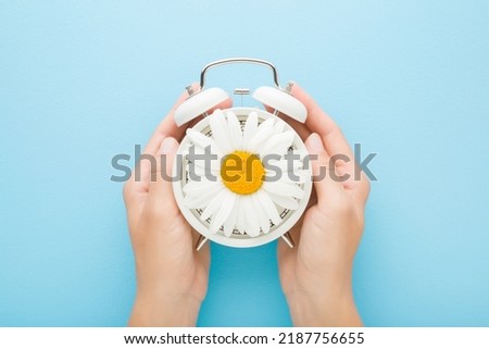 Young adult woman hands holding white alarm clock with big beautiful daisy flower on light blue table background. Pastel color. Time concept. Closeup. Point of view shot. Royalty-Free Stock Photo #2187756655