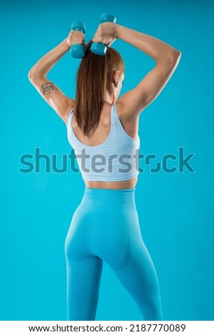 Athlete girl posing from behind while working the triceps in blue clothes isolated on a blue background. Bodybuilding concept.