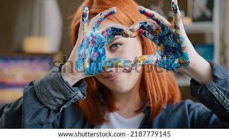 Portrait red-haired girl talented artist female painter stands posing in workshop shows gesture of kindness inspiration love for art sign heart shape with dirty hands in oil acrylic paints smiling
