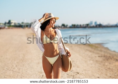 people, summer holidays and leisure concept - happy young woman in bikini swimsuit, white shirt and straw hat with bag walking along beach Royalty-Free Stock Photo #2187785153