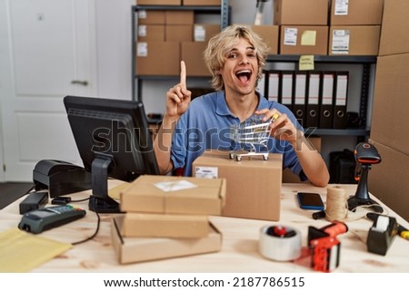Young man working at small business ecommerce holding small cart smiling with an idea or question pointing finger with happy face, number one 